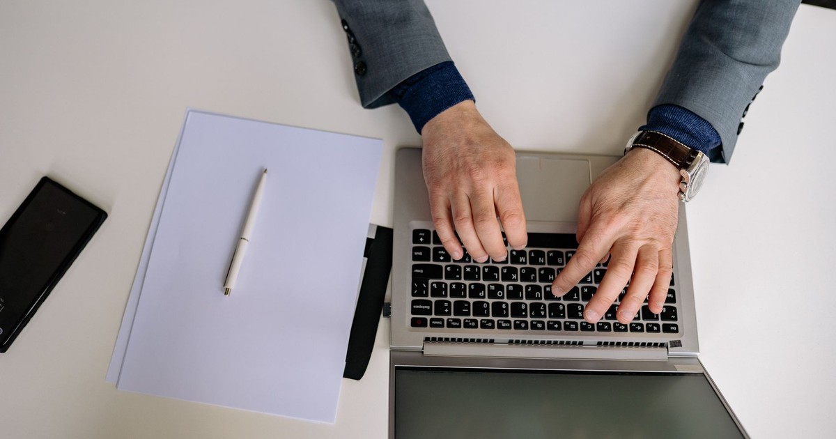 Overhead shot of a man in a suit typing on a laptop with a notepad and pen nearby.