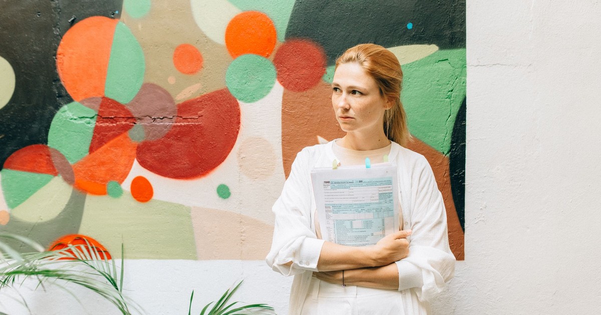 A woman in a white outfit standing indoors holding documents, with a colorful abstract mural in the background.