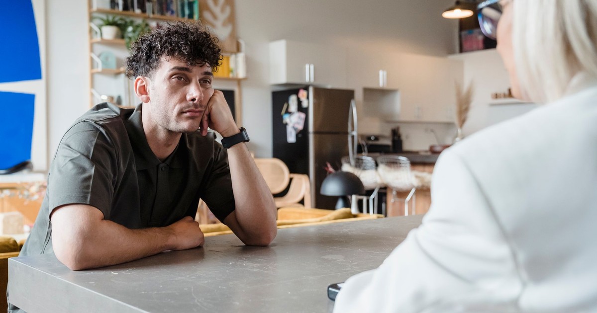 A man with curly hair sits thoughtfully in a modern living room, gazing at a person across the table.