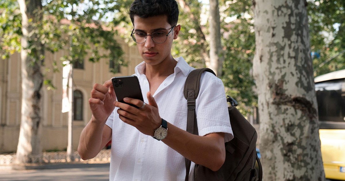 Young man using smartphone on a city street, wearing eyeglasses and a backpack.