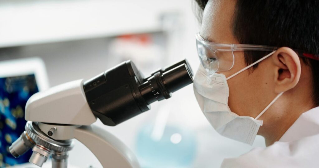 Scientist wearing face mask and goggles examining samples through a microscope in a lab.