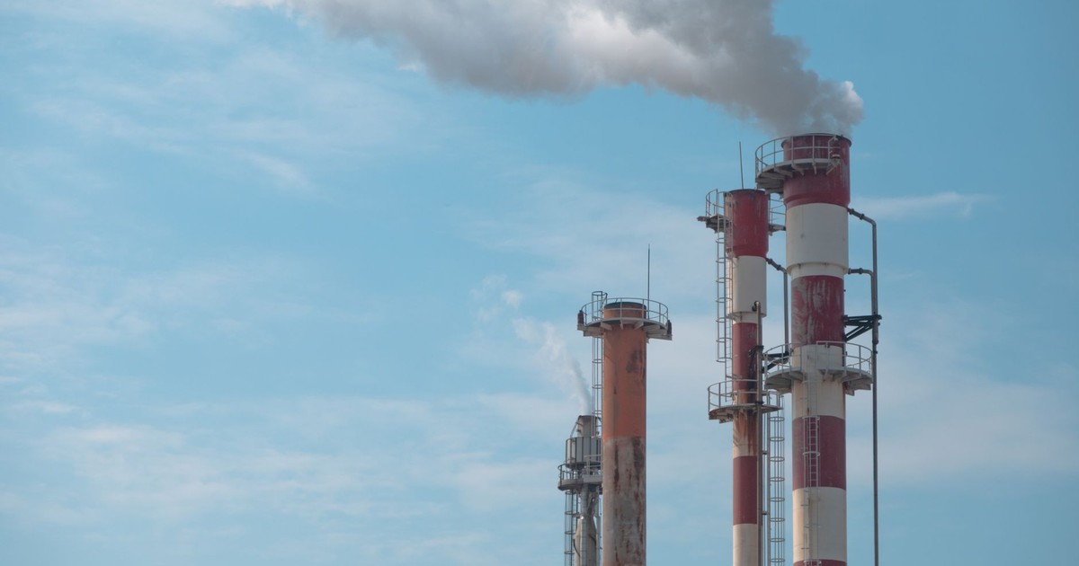 Three industrial smokestacks with white smoke against a clear blue sky, symbolizing air pollution.