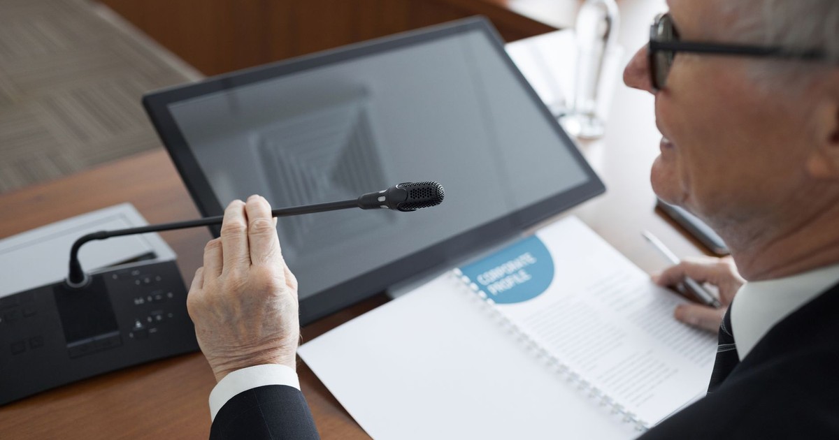 A senior businessman using a microphone during a conference meeting with documents and a monitor.