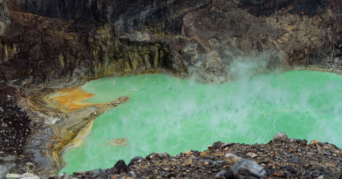 A steaming green acidic lake in a volcanic crater with vibrant color contrast against rocky terrain.