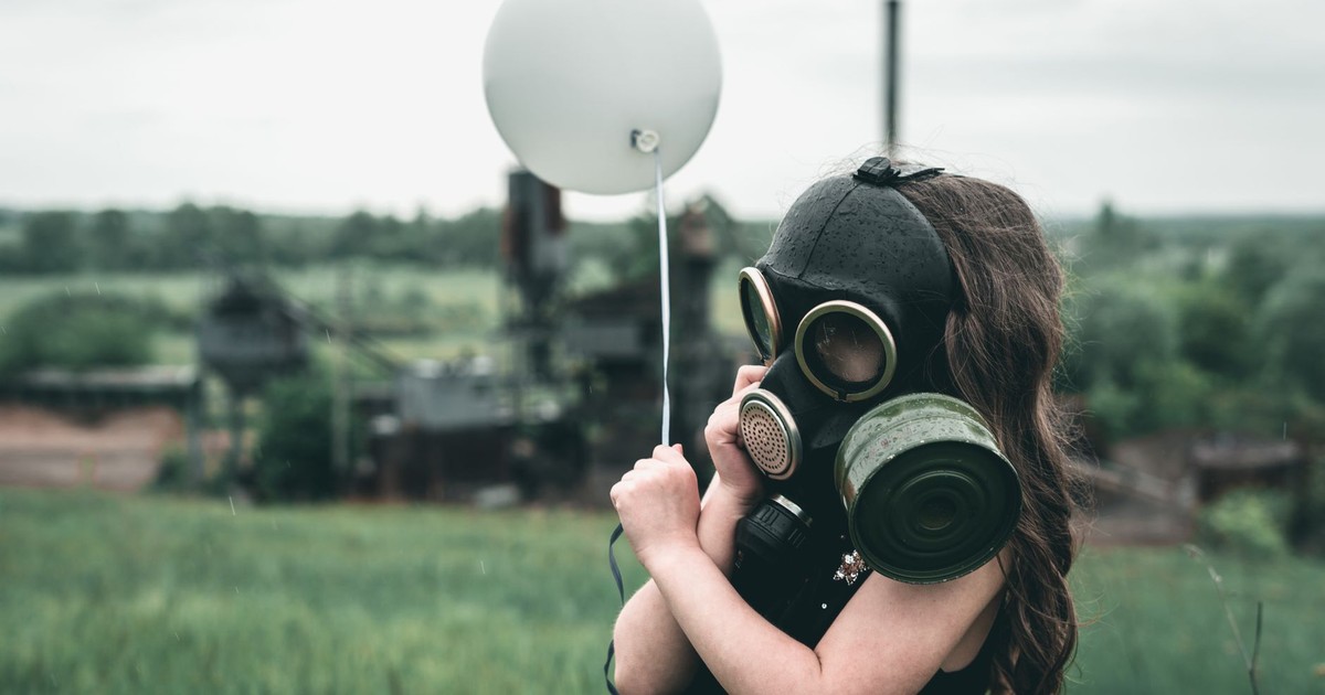 A child wearing a gas mask holds a balloon in a field, symbolizing innocence amidst industrial backdrop.