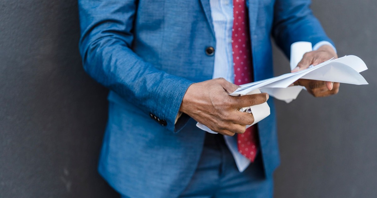 Crop unrecognizable male entrepreneur in formal suit reading documents in hands while standing on gray background in city after workday