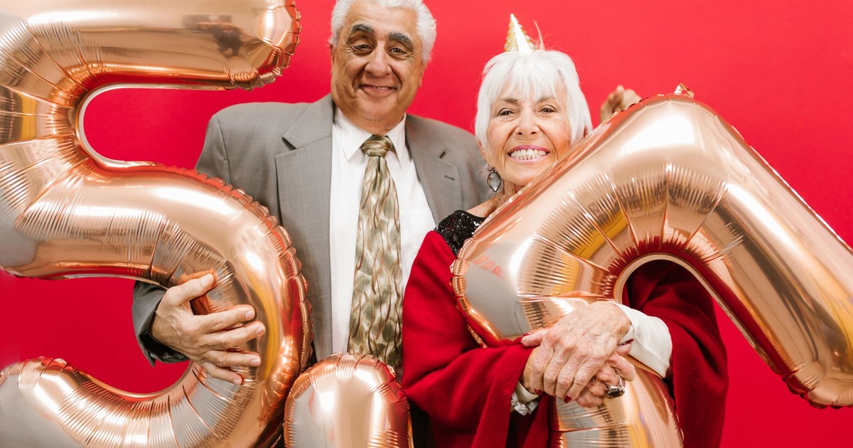 Cheerful senior couple celebrating with gold balloons on a vibrant red background.