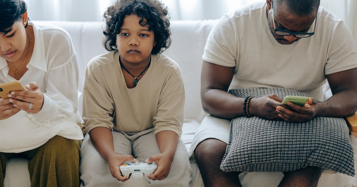 Focused ethnic child playing video game with joystick sitting on sofa between divers parents browsing smartphones