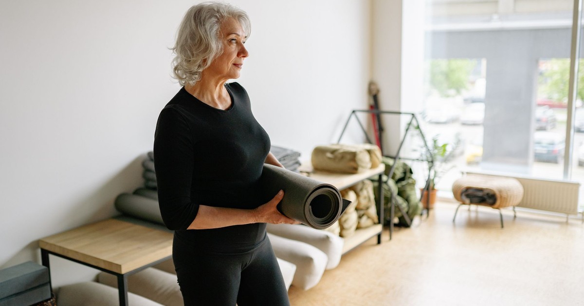 Elderly woman in black attire holding a yoga mat in a bright, minimalist space.