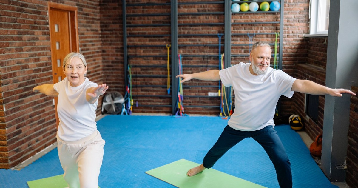 Elderly couple doing yoga poses on mats in a gym, promoting active lifestyle.