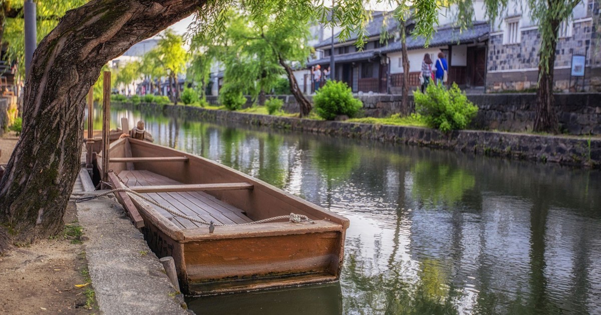 Scenic view of a traditional wooden boat docked along the canal in Kurashiki, Japan, surrounded by lush greenery.