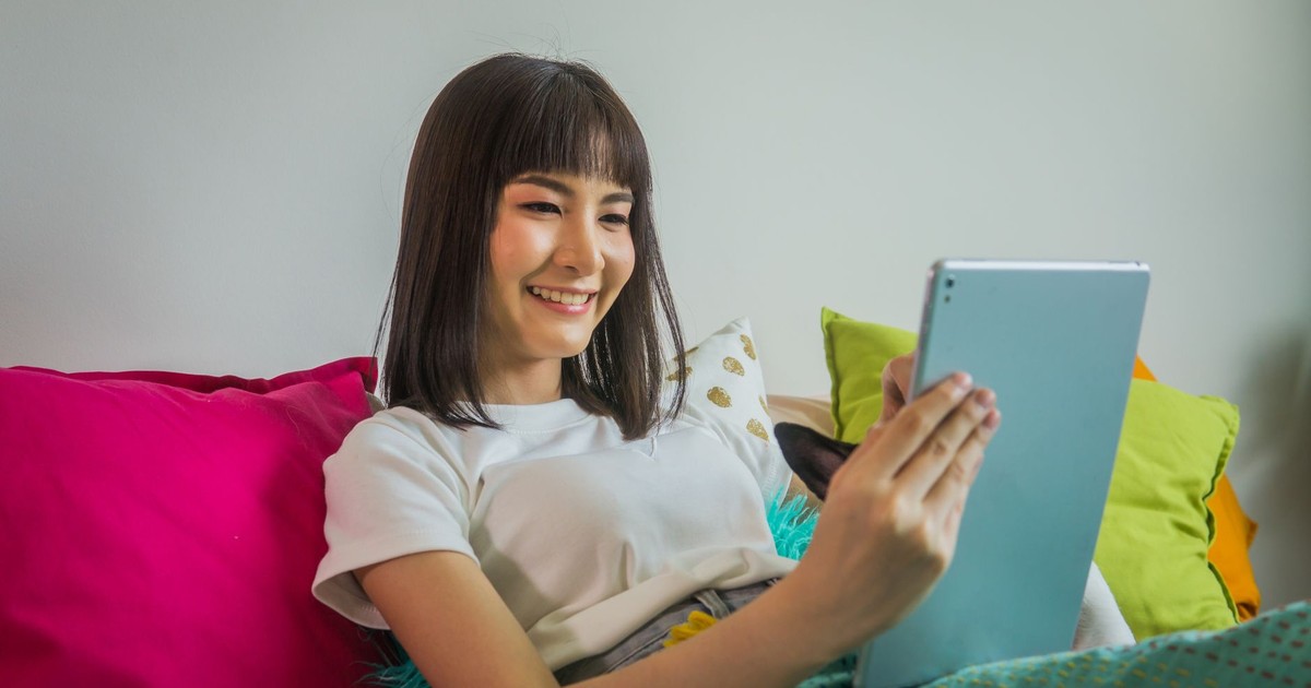 Asian woman with bangs smiling while using a tablet on a couch with colorful pillows indoors.