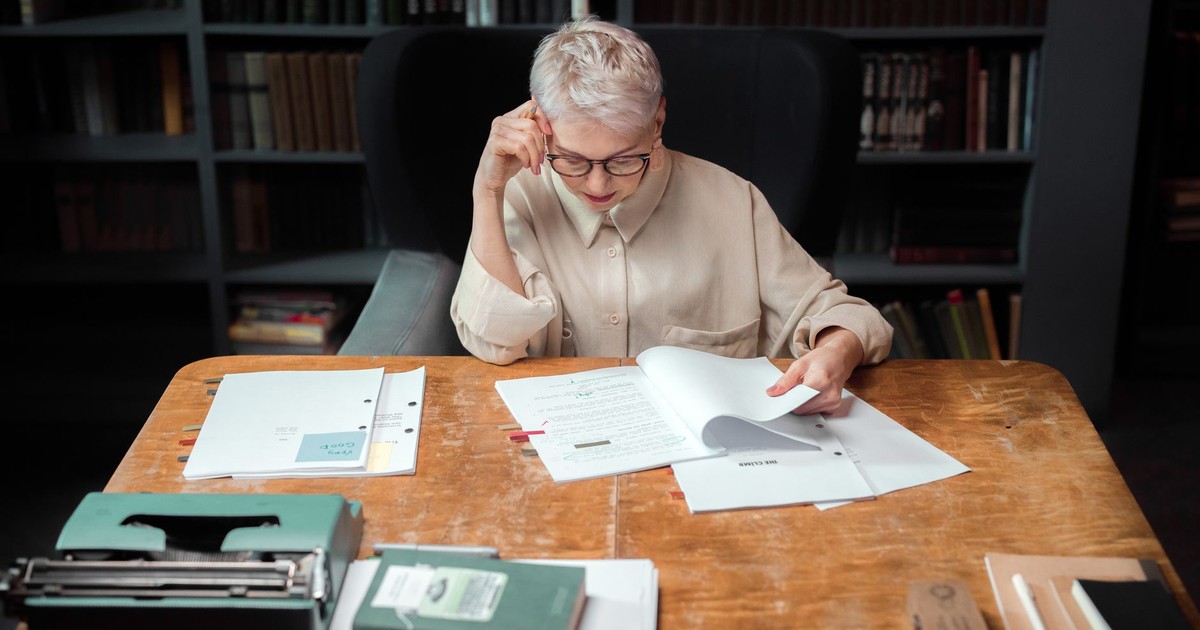 Elderly woman in an office reading and reviewing scripts with a typewriter nearby.
