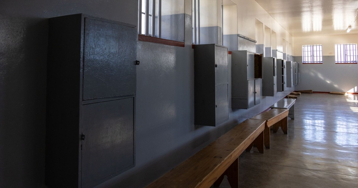 Empty prison cell block with benches and cabinets, lit by barred windows casting shadows.