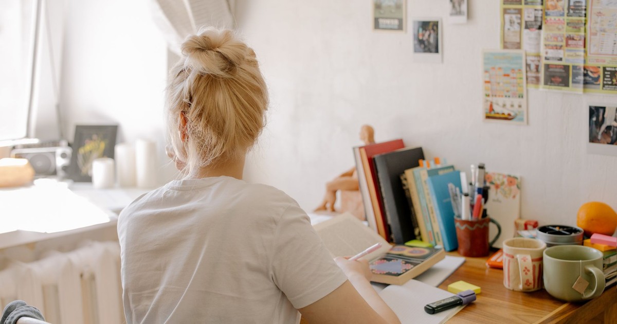 A student with blonde hair studies at a desk in a cozy dorm room setting, surrounded by books and stationery.