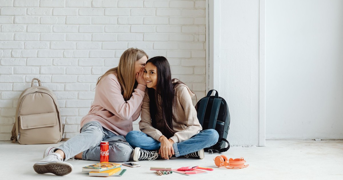Two teenage girls sitting on the floor, whispering and smiling together.