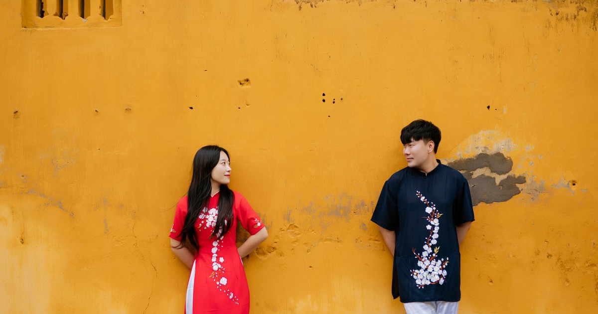 A couple in traditional Ao Dai apparel stand against a bright yellow wall, exuding cultural charm and affection.