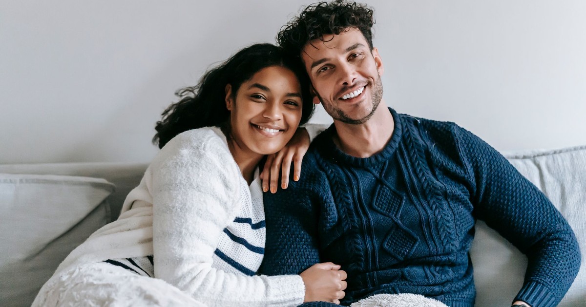 Happy ethnic couple in casual clothes sitting on soft sofa in light room and looking at camera in daytime