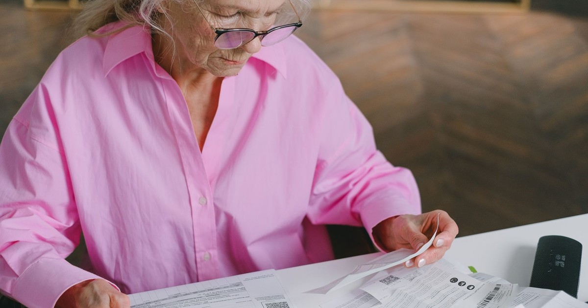 Elderly woman in pink blouse reading documents at a table indoors.