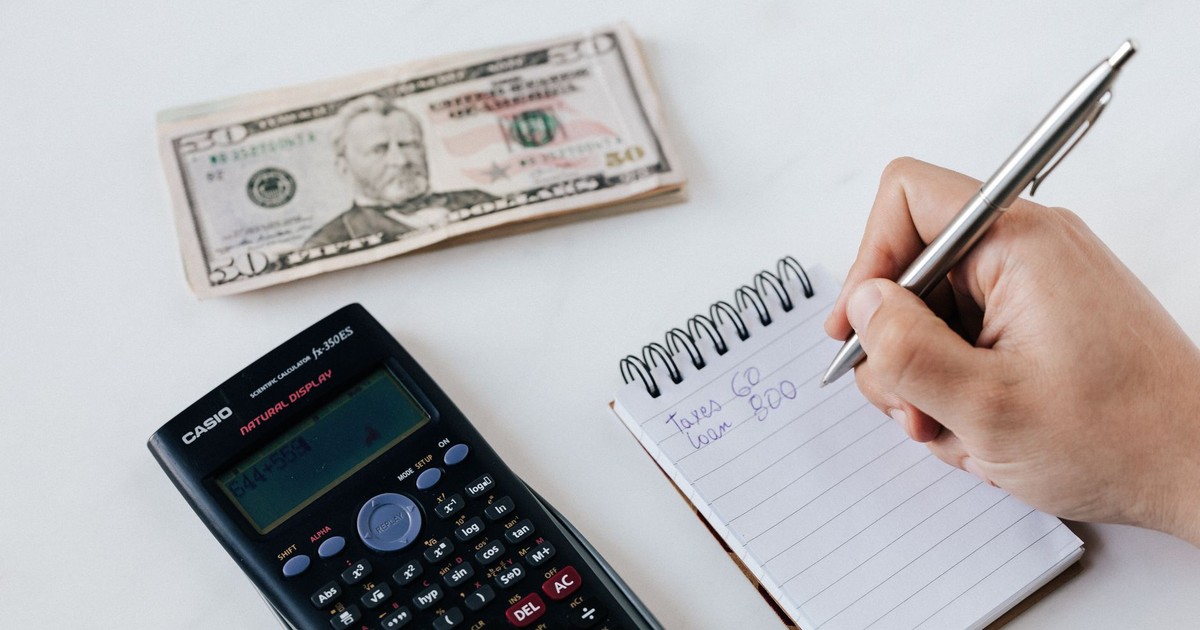 Top view of crop faceless accountant using pen and notepad while counting expenses on table near pile of paper money and calculator