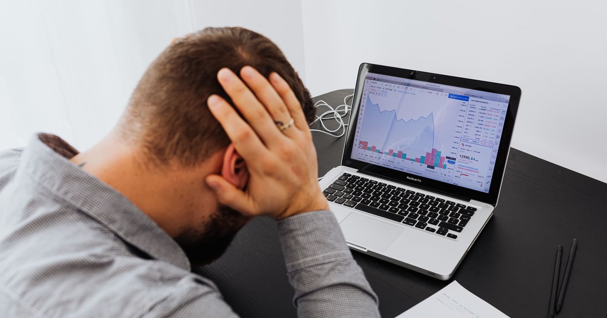 Stressed man at desk looking at declining stock charts on laptop, indicating financial loss.