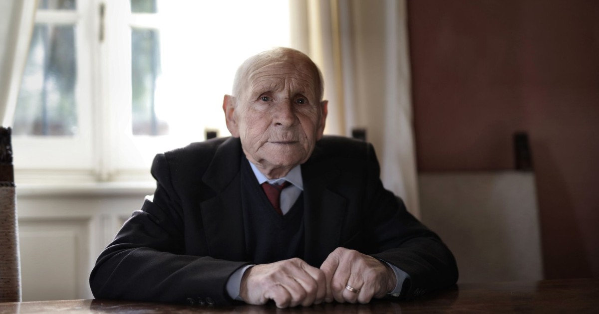A serious elderly man in formal wear sitting at a table indoors, captured during the day.