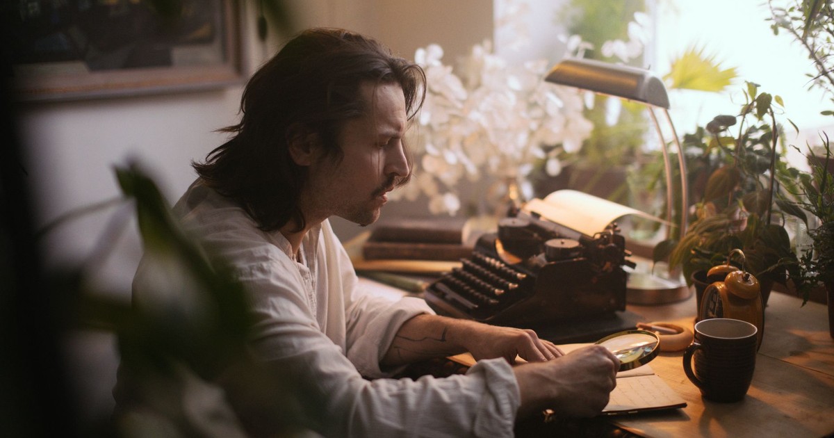 A man with long hair examines a manuscript using a magnifying glass in a cozy study.