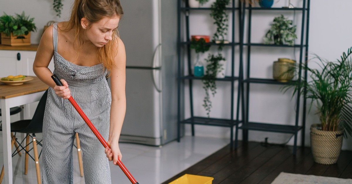 Young woman in jumpsuit with mop leaning forward while washing parquet against potted plants at home