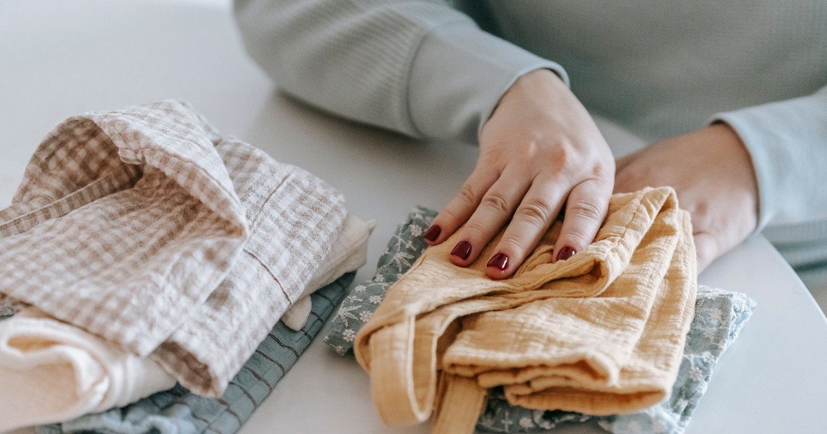 Crop faceless female with red manicure folding colorful baby garments while sitting at white table in light room at home