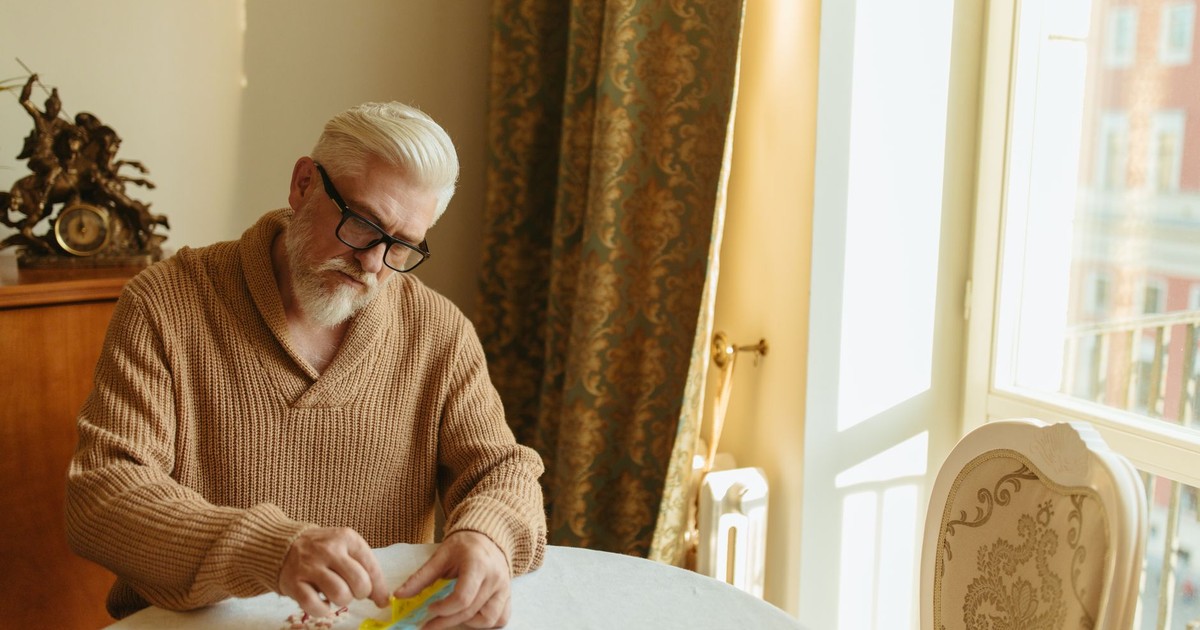 Senior man with gray hair organizing pills at a table by a window, emphasizing daily health routine.
