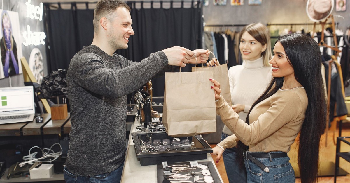 Side view of cheerful male shop assistant standing at counter desk and giving to positive female customers paper bags with purchases in trendy shop