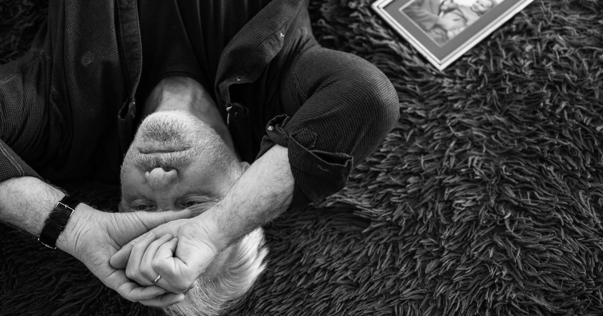 Black and white image of an elderly man lying down, expressing sorrow with a framed photo beside him.