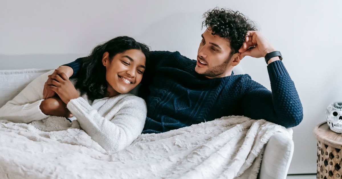 Cheerful young multiethnic couple in casual clothes cuddling and smiling while relaxing on comfortable sofa under warm blanket