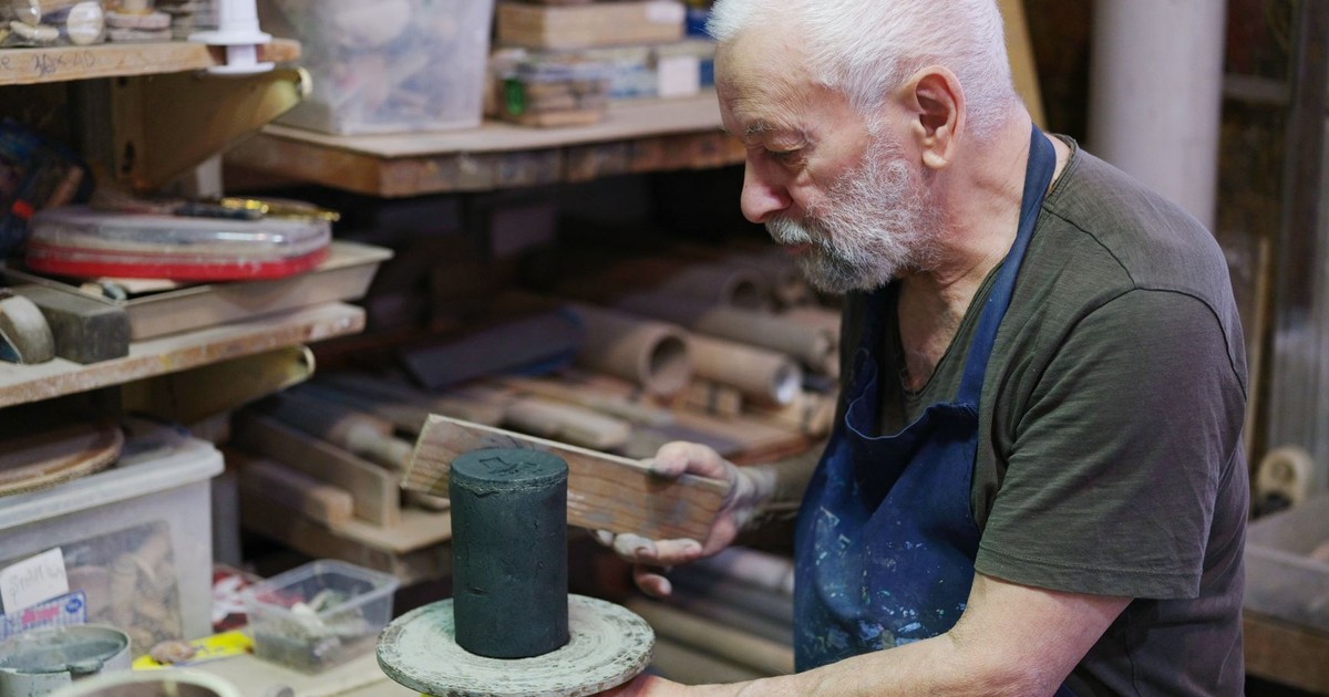An elderly man focuses on crafting in a cluttered workshop, highlighting skilled craftsmanship.