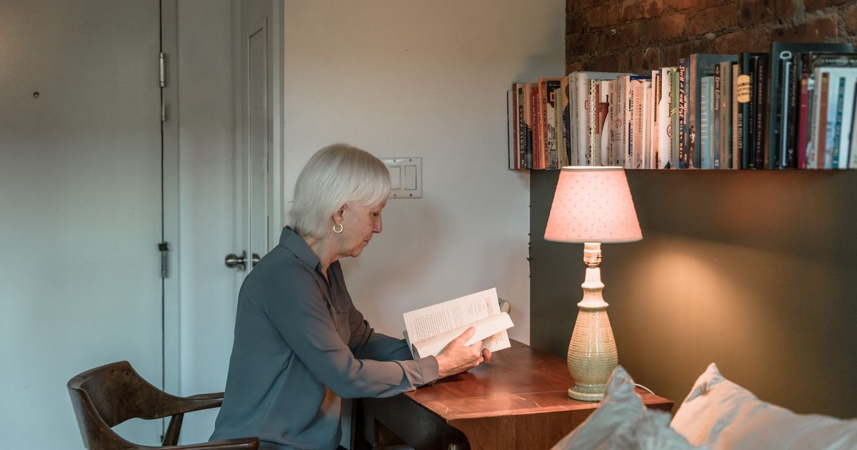 Elderly woman reading a book by a lamp in a cozy home setting.