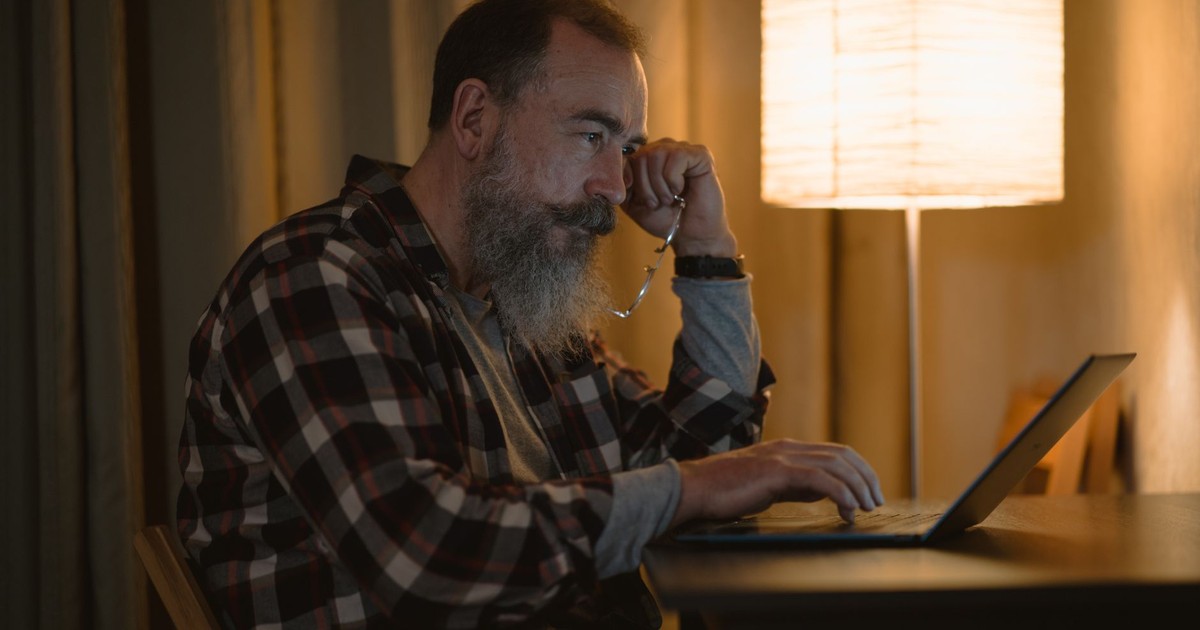 Elderly man with a beard working late on laptop under warm lamp light indoors.