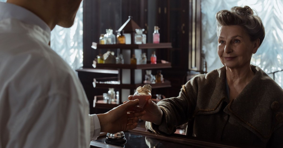 A senior woman receives a perfume bottle from a pharmacist inside a vintage pharmacy.