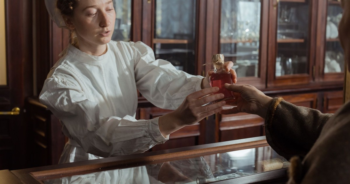 A woman in vintage attire hands a red liquid bottle over a counter, set in a historical apothecary.