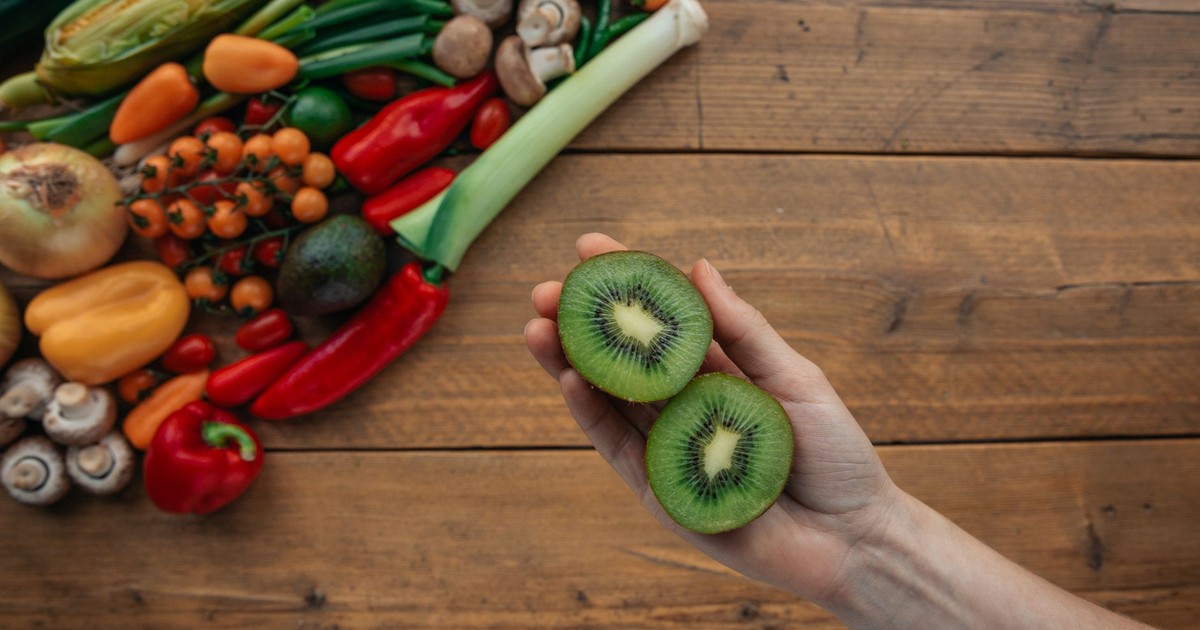 A hand holds sliced kiwi above a wooden table with fresh vegetables, highlighting healthy ingredients.
