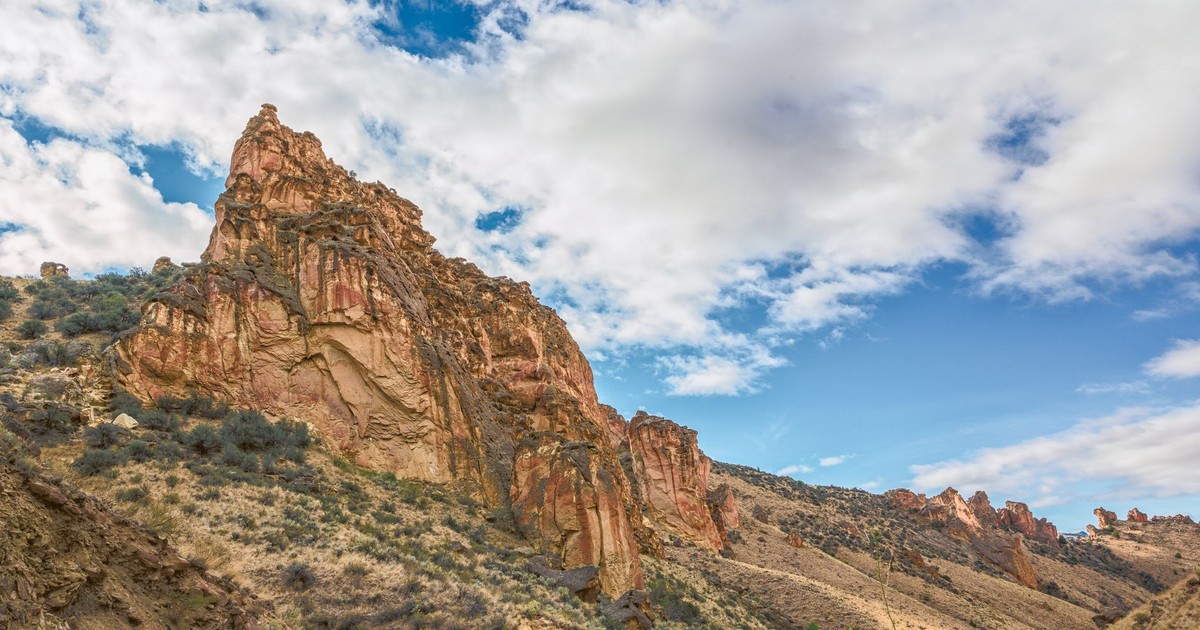 Scenery of rough stony cliff located in mountainous terrain with dry vegetation under blue cloudy sky