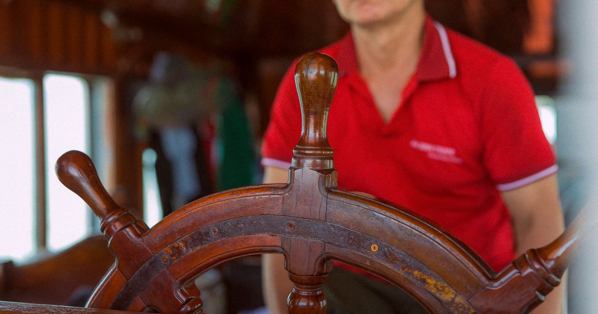 Close-up of a man in red shirt at a ship's wooden helm, steering indoors.