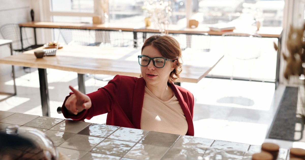 A woman wearing glasses in a red blazer seen ordering in a bright café setting.