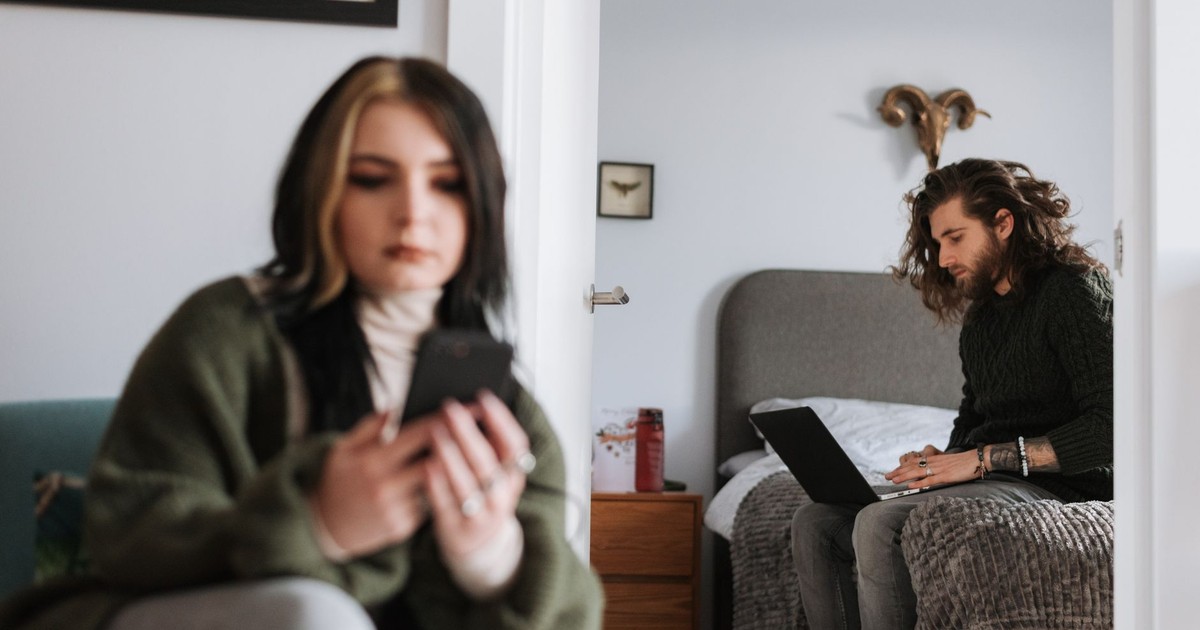 Young hipster male surfing internet on netbook on bed against female partner with cellphone in house