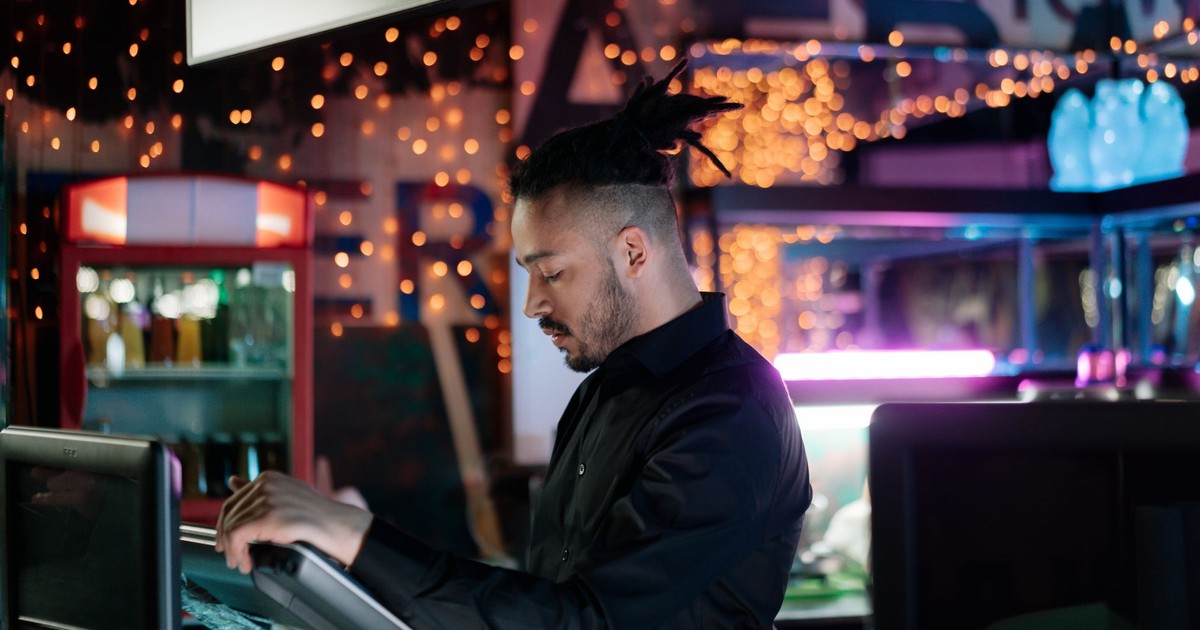 A man with facial hair operates a computer in a trendy bar with festive lights.