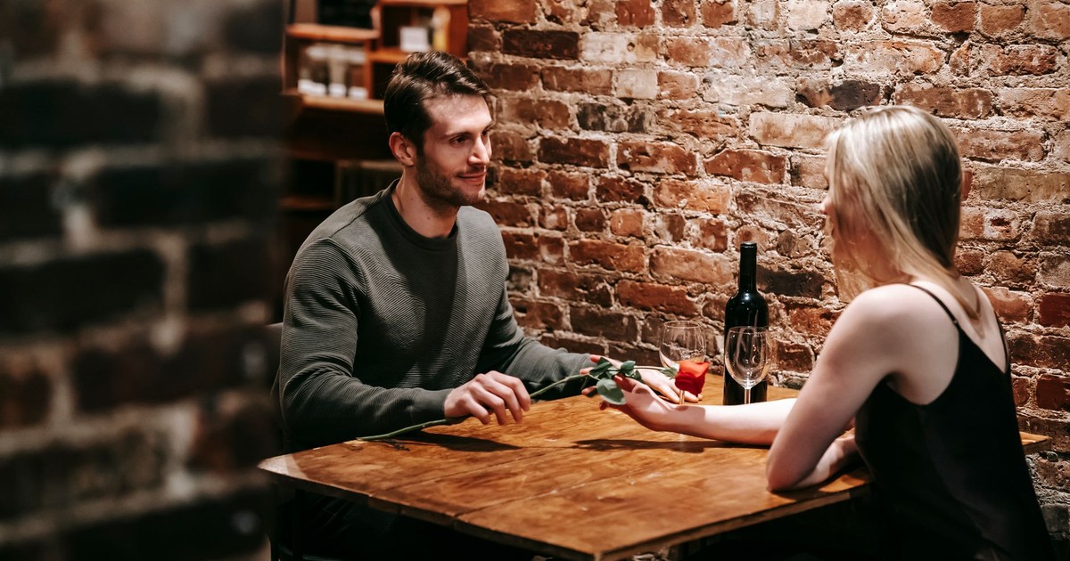Loving boyfriend giving red rose to girlfriend while sitting near brick wall at wooden table during celebration of event in restaurant