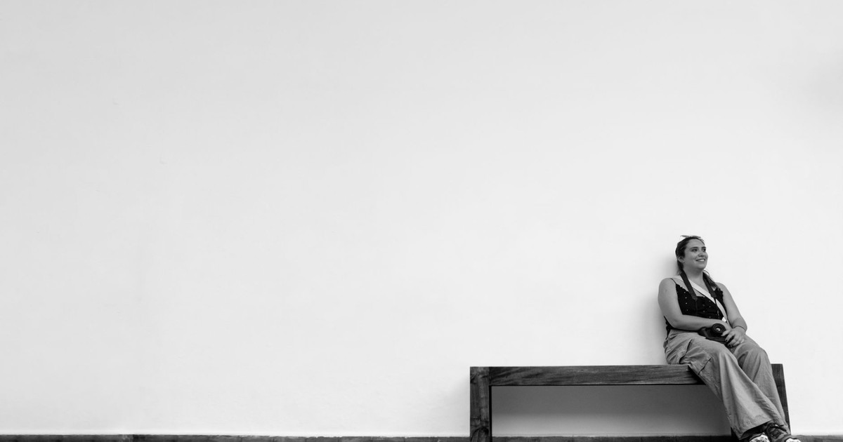Black and white image of woman sitting on bench against a large empty wall, offering ample copyspace.