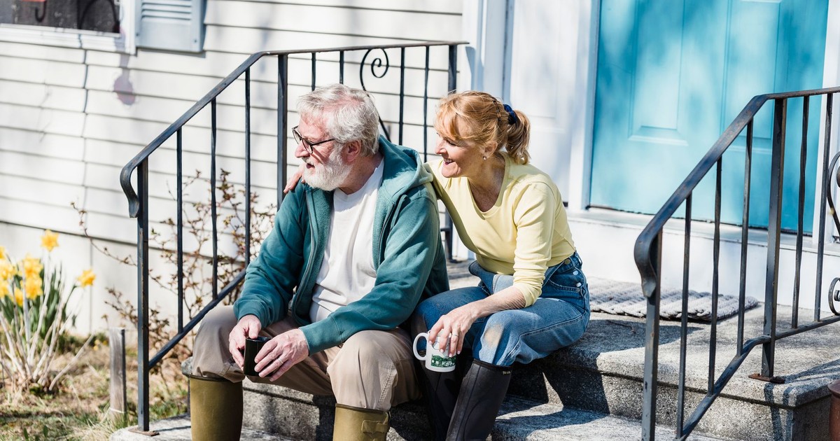 Delightful aged couple in rain boots drinking tea and looking away while sitting on stairs of house in countryside