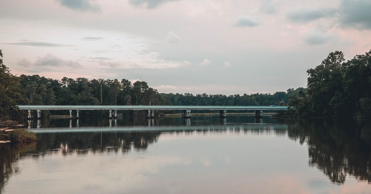 A tranquil scene of a bridge reflecting over a calm river surrounded by trees under a cloudy sky.