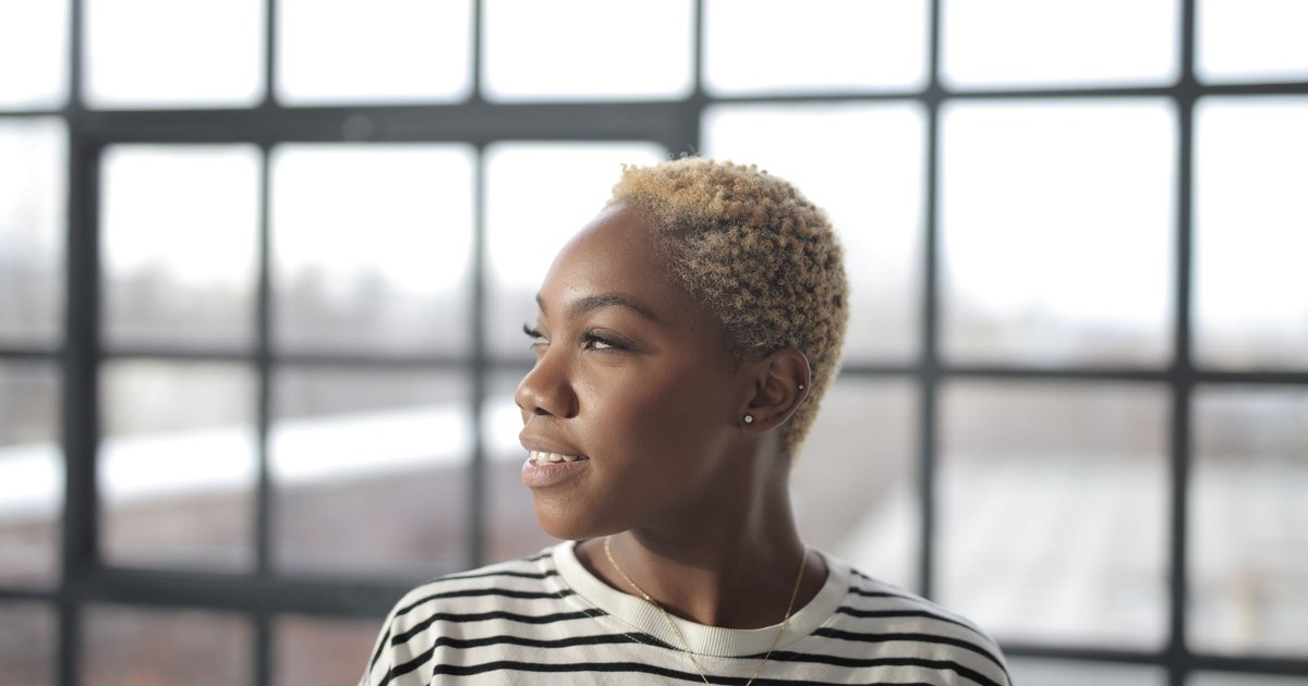 A young woman with short hair gazes thoughtfully out of a window, framed by soft natural light indoors.