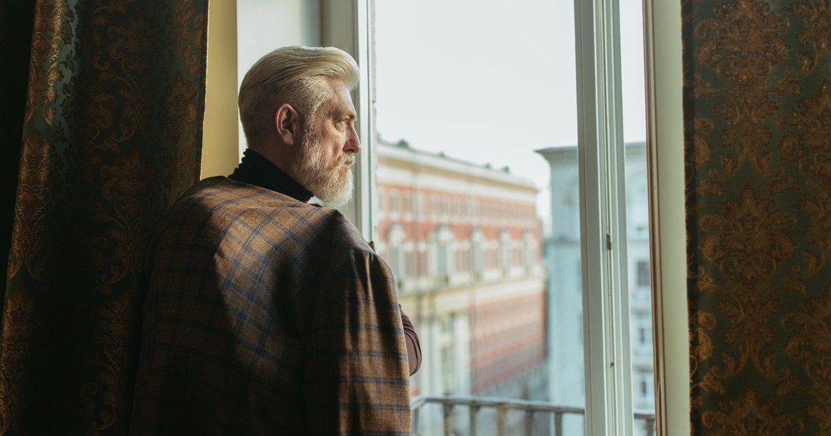 Elderly man with a beard gazing out a window in a classic interior setting.